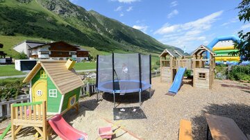 A beautiful playground with a green playhouse, a slide, a trampoline, and a bouncy castle in the background. The surroundings are green and mountainous under a blue sky.