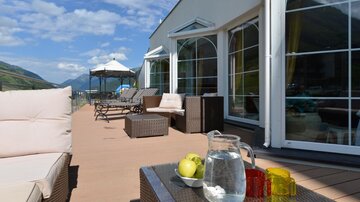 An inviting balcony with cozy furniture and a table on which a jug of water and apples are placed. In the background, mountain landscapes and a clear sky are visible.