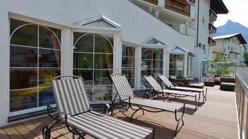 A terrace with loungers and large windows of a house. Mountains can be seen in the background.