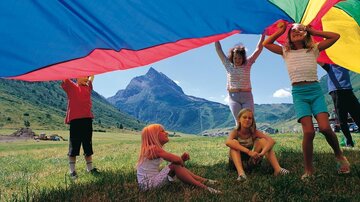 A group of children is playing under a colorful cloth in nature. In the background, green meadows and mountains can be seen.