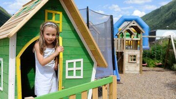 A girl is standing in a colorful playhouse on a playground. In the background, there are more play equipment and a beautiful landscape visible.