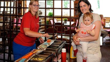 A woman with glasses serves hot dogs in a restaurant. Another woman holds a small child in her arms while smiling.