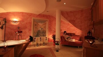 A relaxing bathroom with a bathtub and soft lighting. One person is relaxing in the bath while another is reading on a sofa.