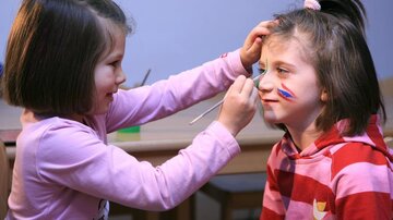 Two girls are playing and painting each other's faces. One girl has colorful paint on her cheeks.