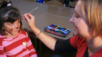 A child in a striped sweater is receiving face paint from an adult. On the table, there is a palette with various colors.