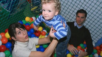 A small boy is standing in a ball pit while a woman lifts him up. A man is sitting next to them and observing the situation.
