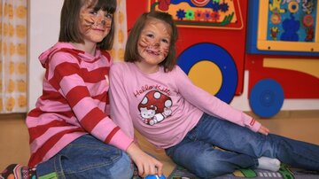 Two children with animal faces are sitting smiling on the floor. They are wearing colorful clothes and are playing in a colorful room.