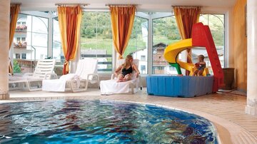 An indoor pool with clear water and a slide for children. In the background, adults are sitting on loungers, and there are large windows overlooking green surroundings.