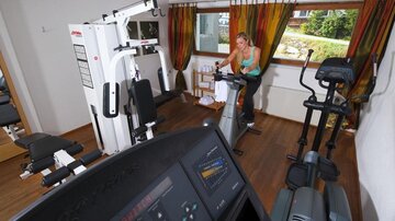 A well-equipped fitness room with various machines. A woman is exercising on a stationary bike.