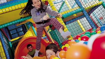 Two children are playing in a colorful play paradise with a slide and many balls. A girl is happily jumping in the air, while a boy in the background is laughing and playing in the ball pit.