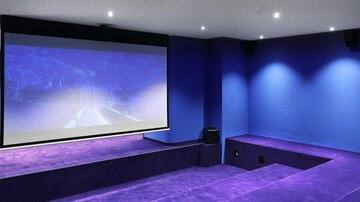 A modern home theater room with a large projection screen and a purple carpet area. The walls are painted in a deep blue, and the ceiling has dimmable lights.