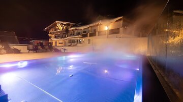 A illuminated pool at night, surrounded by a steaming atmosphere. In the background, a stylish building is visible.