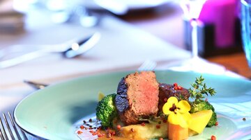 An artfully arranged plate with tender meat, broccoli, and colorful vegetables. The presentation is elegant and appealing, with an edible flower as decoration.