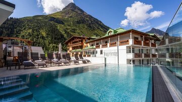 A modern hotel with a swimming pool and loungers in the foreground. In the background, green mountains and a blue sky can be seen.