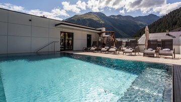 A beautiful pool with clear water and loungers. In the background, there are mountains and a blue sky.