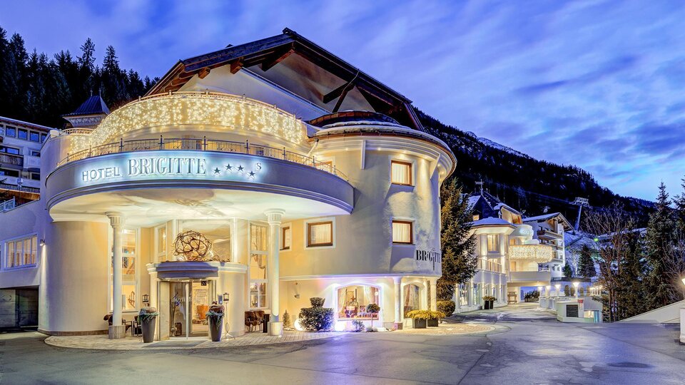 Ein elegantes Hotelgebäude mit einer einladenden Fassade und warmen Lichtern. Die Umgebung zeigt eine malerische Berglandschaft im Hintergrund.