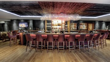 Eine moderne Bar mit einem geschwungenen Thekenbereich und stilvollen, hochmodernen Barhockern. Der Raum hat eine warme Atmosphäre mit Holzfußboden und indirekter Beleuchtung.