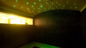 Ein entspannender Raum mit dunklen Wänden und einem funkelnden Sternenhimmel in grünen Lichtern. Sanftes gelbes Licht strömt durch ein Fenster.