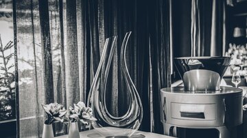 Ein eleganter Tisch in einem Restaurant mit einer stilvollen Blumenvase und zwei kleinen Blumenarrangements. Im Hintergrund sind Vorhänge und ein modernes Dekor zu sehen. | © elizabeth.at