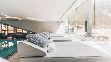 Ein moderner Lounge-Bereich mit gemütlichen Liegen und großen Fenstern. Der Blick aus dem Raum zeigt eine ruhige Umgebung mit einem Pool. | © elizabeth.at