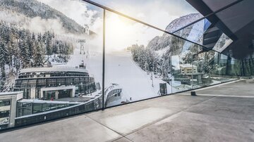 Eine moderne Architektur mit großen Fenstern, die einen Blick auf schneebedeckte Berge und eine Skipiste bieten. Die Szene vermittelt eine winterliche Atmosphäre. | © elizabeth.at
