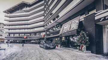 Ein modernes Gebäude mit mehreren Etagen in einer verschneiten Umgebung. Vor dem Eingang steht ein elegantes Auto, und Weihnachtsdekorationen sind sichtbar. | © elizabeth.at