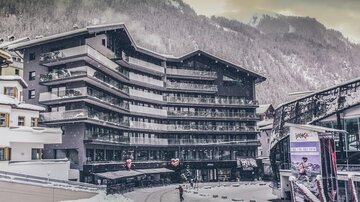 Ein modernes Hotel in einem verschneiten Bergdorf. Im Hintergrund sind schneebedeckte Berge und eine ruhige Umgebung zu sehen. | © elizabeth.at
