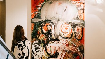Eine Person steht auf einer Treppe und betrachtet ein großes, abstraktes Gemälde an der Wand. Das Kunstwerk zeigt kräftige Farben und einen auffälligen Gesichtsausdruck.