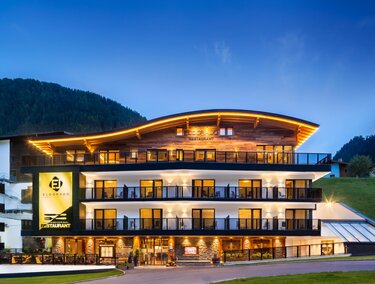 Ein modernes Hotelgebäude mit beleuchtetem Dach und großen Balkonen. Die Umgebung ist von Hügeln und einer klaren Abendatmosphäre geprägt.