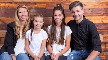 Eine Familie posiert vor einer Holzverkleidung. Zwei Mädchen sitzen zwischen ihren Eltern, alle lächeln freundlich.