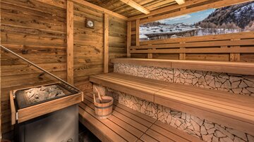 Eine gemütliche Sauna mit hölzernen Bänken und einem Steinofen. Durch das Fenster ist eine winterliche Berglandschaft sichtbar.