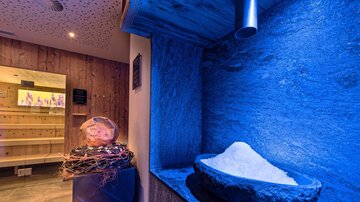 Ein Wellnessbereich mit blauen Lichtern und einem Steinsalzbecken. Im Hintergrund ist eine Holzsauna zu sehen.