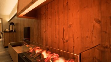 Ein moderner Kamin mit glühenden Holzscheiten in einem stilvollen Raum. Die Holzwände und die sanfte Beleuchtung schaffen eine gemütliche Atmosphäre.