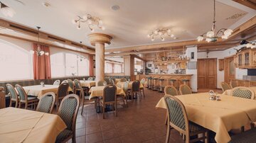 Ein gemütliches Restaurant mit braunen Holzelementen und gelben Tischdecken. Es gibt viele Tische und einen Bereich mit einer Bar im Hintergrund.