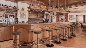 Ein gemütlicher Barbereich mit einem hölzernen Tresen und Barhockern. Im Hintergrund sind Regale mit Getränken und eine einladende Atmosphäre zu sehen.