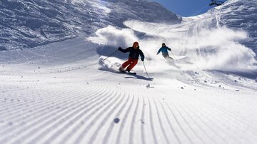 Zwei Skifahrer fahren auf einer schneebedeckten Piste. Die Sonne scheint und der Himmel ist klar.
