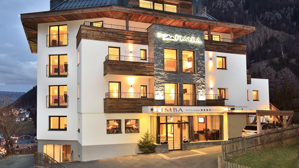 A modern hotel building with multiple windows, balconies, and an inviting entrance. The facade is in neutral colors and is surrounded by a beautiful mountain landscape.