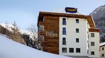 A modern hotel building with a mix of wood and white facade. In the background, snow-capped mountains and a clear sky can be seen.