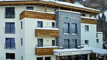 A modern hotel in alpine style with wooden paneling and large windows. The snow covers the entrance and surroundings, while the mountains are visible in the background.