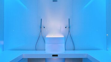 A modern bathroom with blue lighting. In the foreground, there is a stylish toilet and two showers.