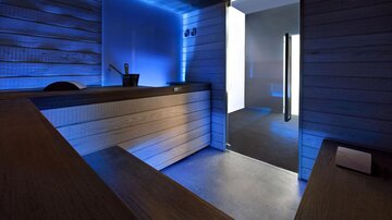 A modern wellness room with wooden paneling and blue lighting. There is a wooden area and access to another room.