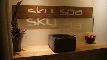 An elegant reception area of the Sky Spa with a wooden sign and a stone washbasin. On the shelf, there are fruits in a bowl.
