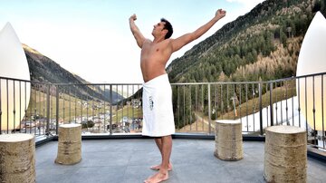 A naked man stands on a terrace with his arms stretched into the air. In the background, there are green mountains and a clear blue sky.