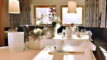 An elegant table in a restaurant with a white tablecloth and decorative flowers. In the background, bright windows and a cozy atmosphere can be seen.