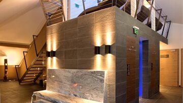 A modern and stylish reception hall with gray stone walls and warm lighting. In the background, stairs and an elegant wooden ceiling can be seen.