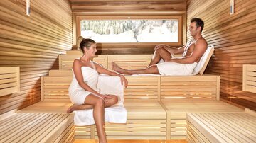 A modern steam room with bright wooden walls. Two people relax in towels on the wooden benches.