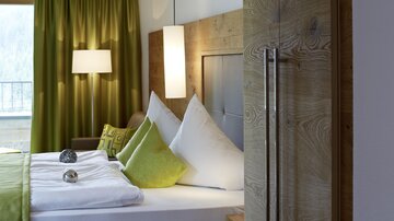 A modern bedroom with a large bed and cozy pillows. The walls are painted in warm colors and there are large windows with curtains.