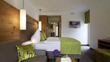 A modern hotel room with a cozy bed and a gray sofa. The decor is in warm colors, with a touch of green accent.