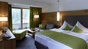 A modern hotel room with a large bed, comfortable furniture, and a balcony. The green accents and warm wood details create a cozy atmosphere.