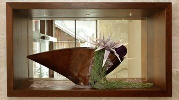 An elegant wooden sculpture in a display case, adorned with decorative plants and flowers. The background features a stylish setting with windows and natural light.